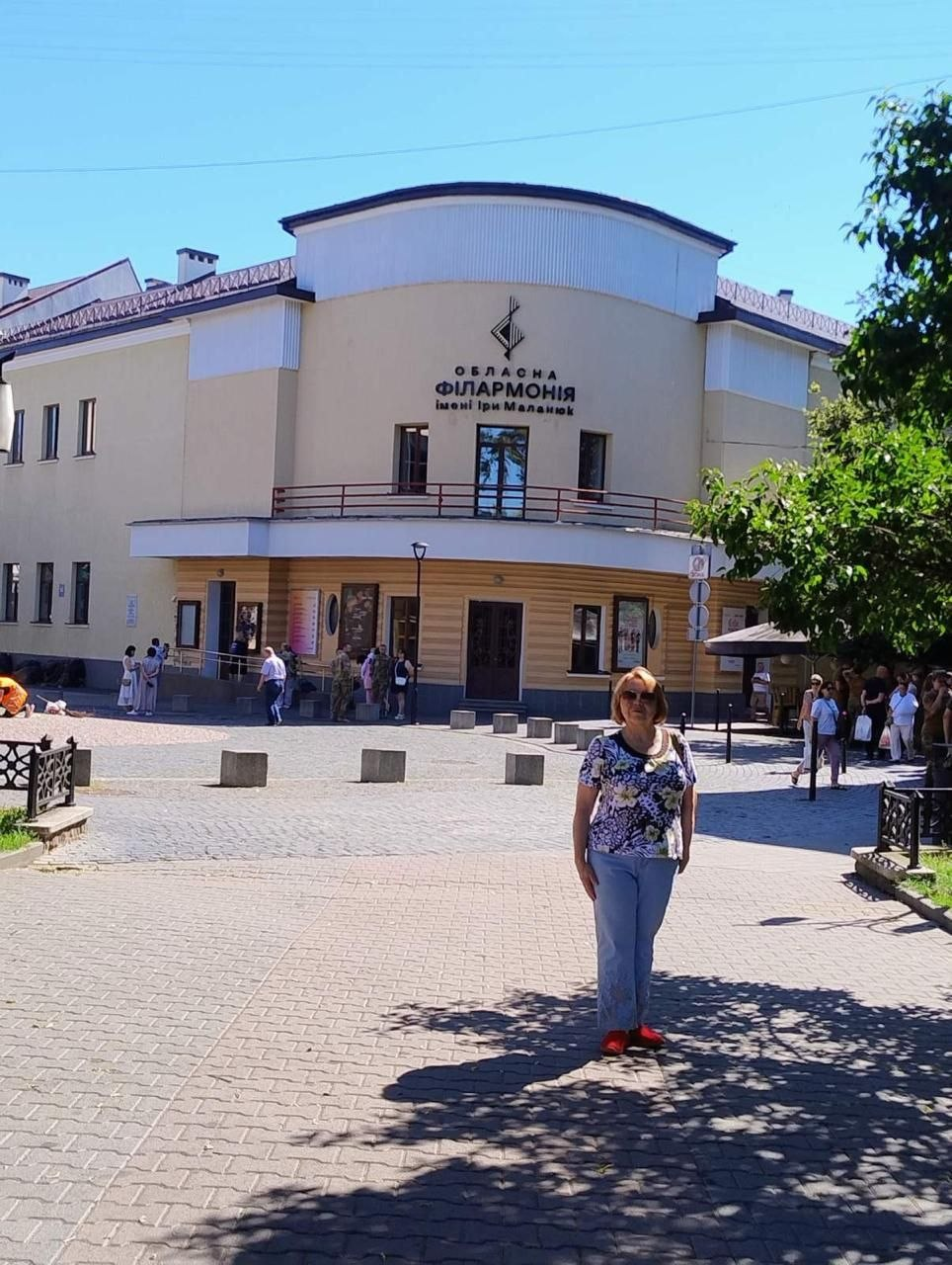 Жінка стоїть на брукованій площі на тлі двоповерхової будівлі Обласної філармонії імені Ірини Маланюк. На світлому фасаді будівлі розміщено логотип і назву закладу, а навколо видно перехожих та зелені дерева в сонячний день.
