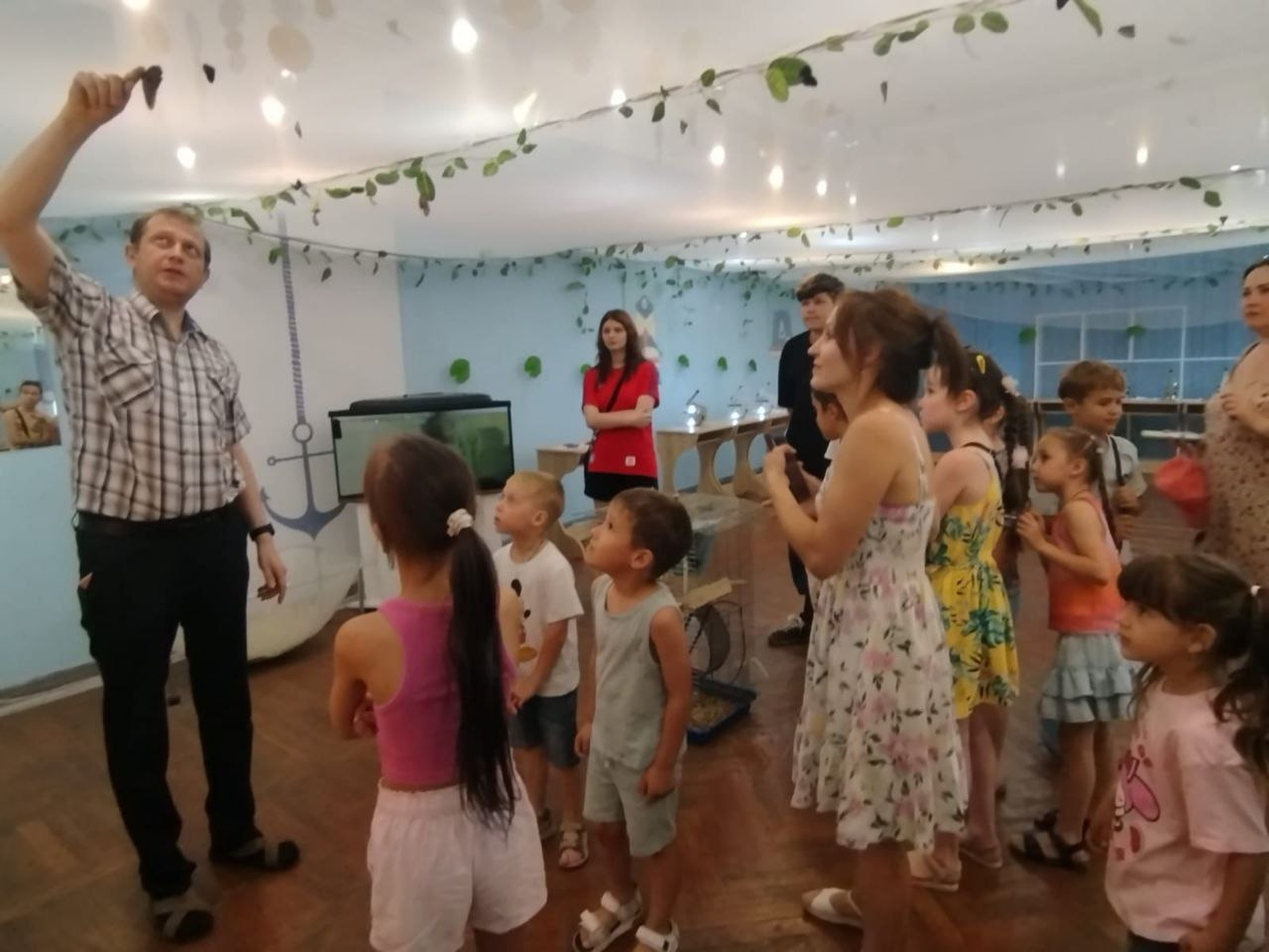 Чоловік у сорочці в клітинку показує рукою вгору, проводячи екскурсію для групи дітей та дорослих у світлій залі. Відвідувачі уважно слухають і дивляться в напрямку його руки, а приміщення прикрашене штучними зеленими гірляндами під стелею.