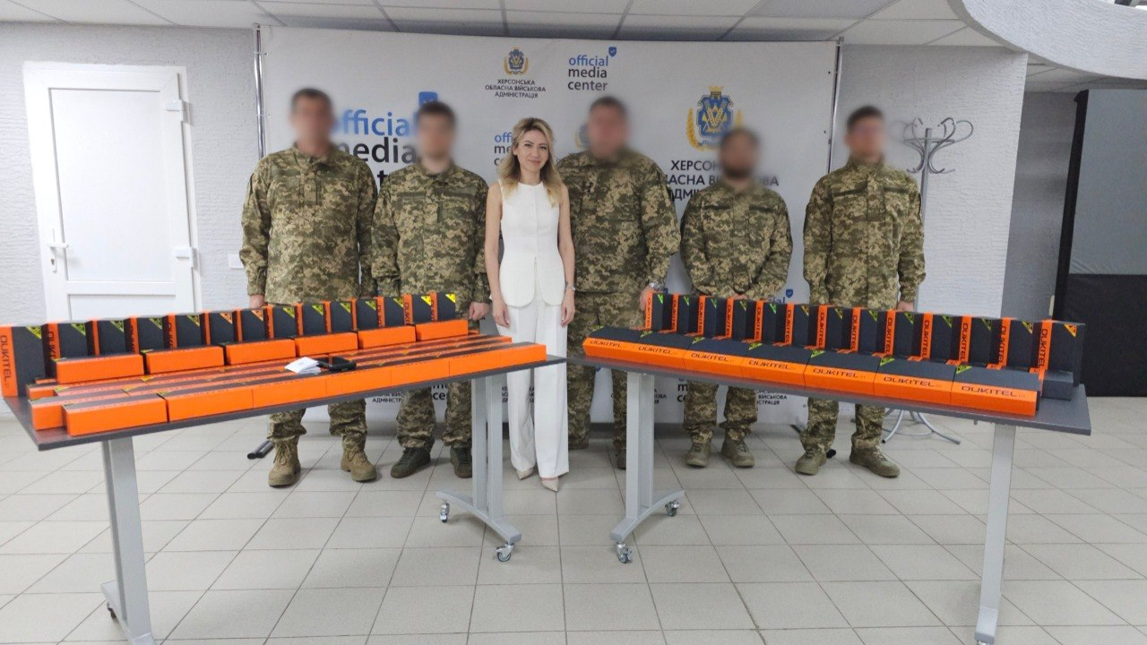 Жінка у білому костюмі та п'ятеро військовослужбовців у камуфляжній формі із розмитими обличчями стоять у приміщенні на тлі банера «official media center Херсонська обласна військова адміністрація». Перед ними на двох столах розкладено велику кількість однакових помаранчево-чорних коробок із технікою.