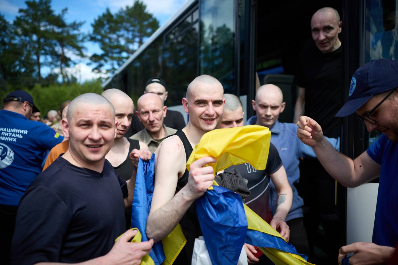 Група чоловіків із поголеними головами виходить з автобуса на відкритому повітрі. Один із чоловіків у центрі посміхається та тримає в руках синьо-жовтий прапор України, інші пасажири також тримають стяг або стоять поруч, а ліворуч видно представника у синій футболці з написом про координаційний штаб.