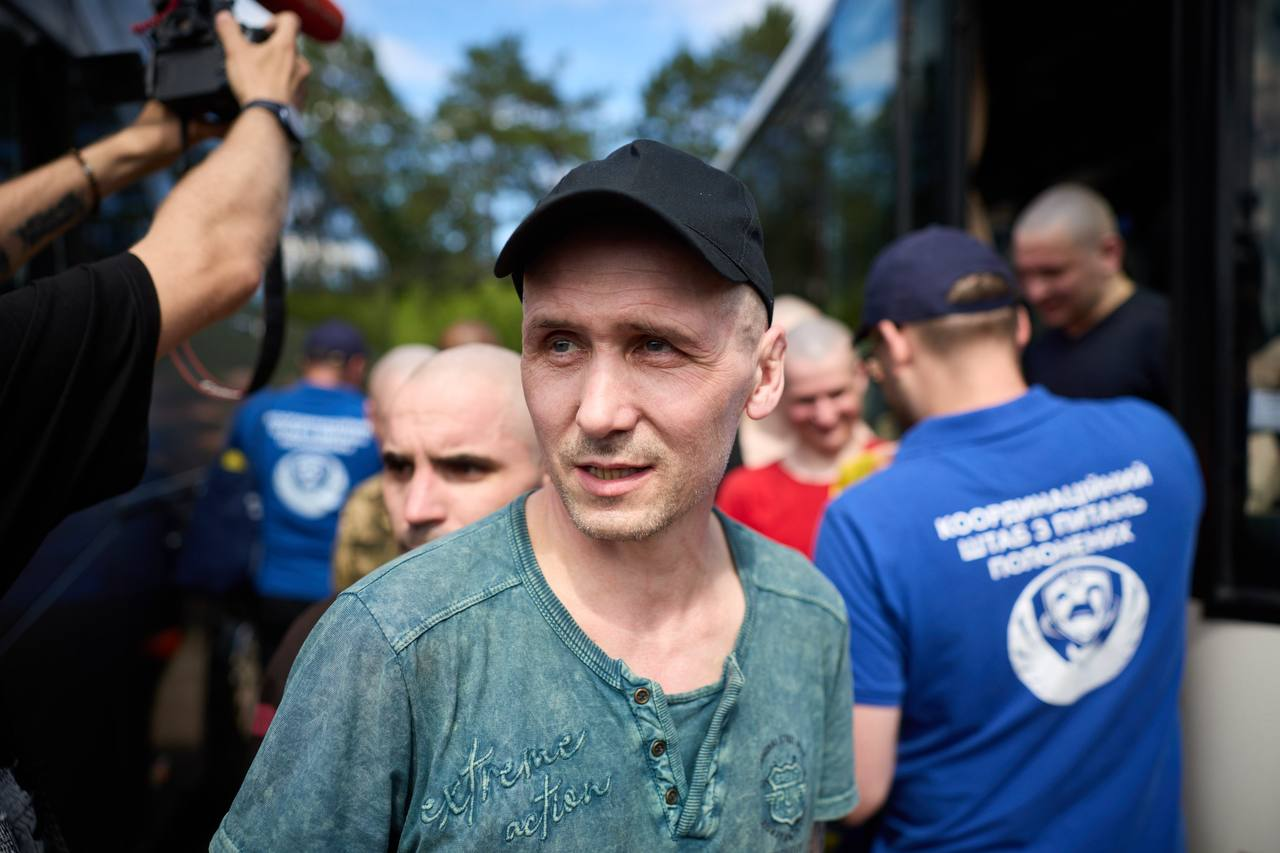 Чоловік у чорній кепці та зеленій футболці стоїть на передньому плані перед автобусом. Праворуч від нього стоїть представник у синій футболці з логотипом та написом «Координаційний штаб з питань поводження з військовополоненими», а ліворуч журналіст тримає відеокамеру.