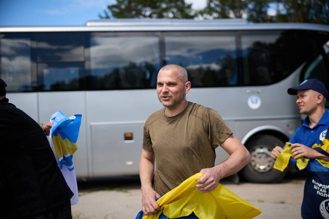 Чоловік у зеленій футболці стоїть на передньому плані на тлі великого сірого автобуса, тримаючи в руках синьо-жовтий прапор. Праворуч від нього стоїть представник у синій формі Координаційного штабу з питань поводження з військовополоненими, який тримає жовту стрічку.