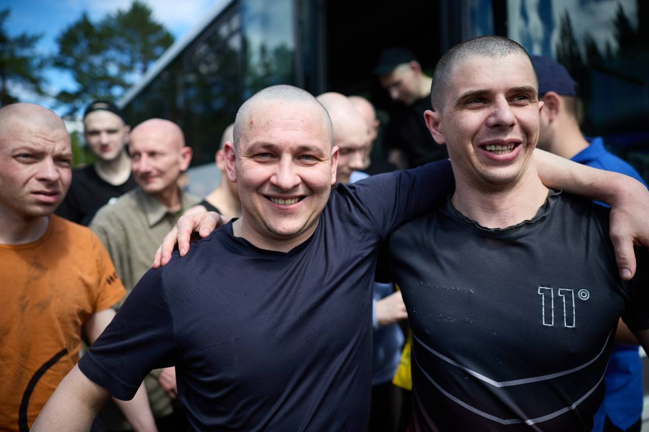 Двоє чоловіків із поголеними головами на передньому плані щиро посміхаються та обіймають один одного за плечі. Вони стоять на тлі великого автобуса, з якого виходять інші чоловіки, а на задньому плані проглядаються зелені дерева та синє небо.