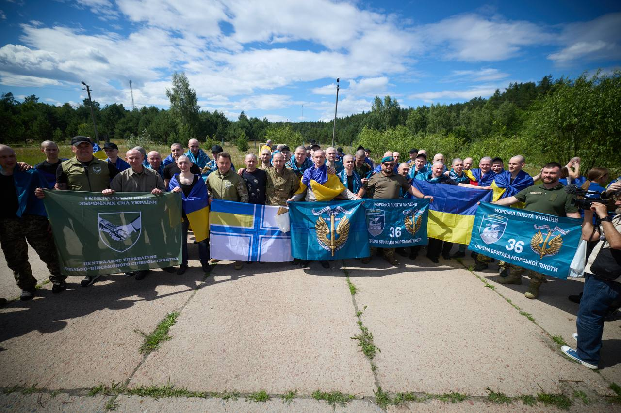 Велика група військовослужбовців та цивільних осіб стоїть у шеренгу на бетонному майданчику просто неба. Вони тримають державні прапори України, прапори 36-ї окремої бригади морської піхоти та банер Центрального управління цивільно-військового співробітництва Генерального штабу ЗСУ, а на задньому плані видно ліс і хмарне небо.