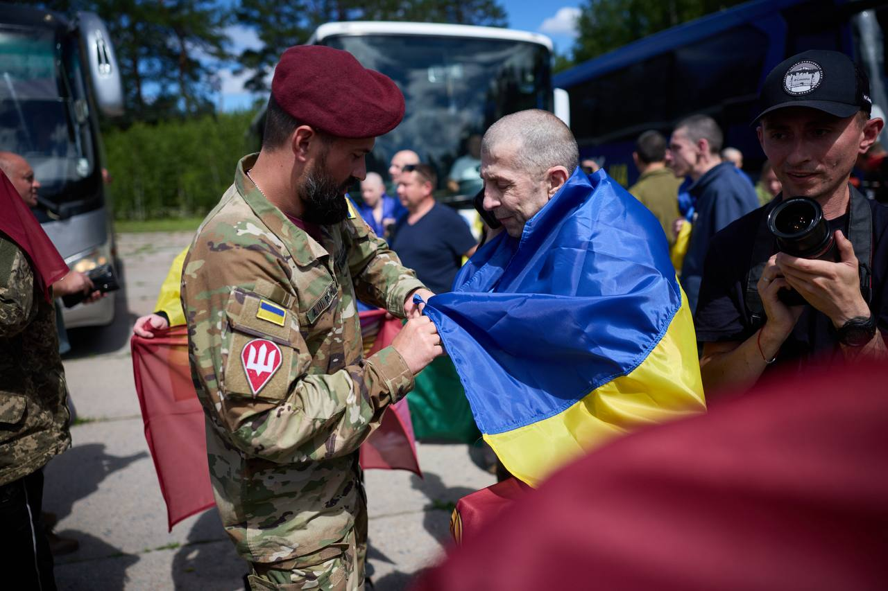 Військовослужбовець у камуфляжній формі та бордовому береті допомагає загорнутися в державний прапор України виснаженому чоловікові на відкритому майданчику. Праворуч стоїть чоловік у чорній кепці з відеокамерою, а на задньому плані проглядаються автобуси, інші люди та зелені дерева.