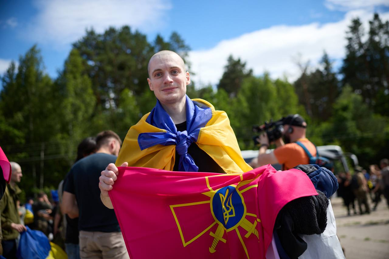 Чоловік із поголеною головою стоїть на передньому плані, накинувши на плечі синьо-жовтий прапор України та тримаючи в руках бордове знамено Збройних Сил України. На розмитому задньому плані видно інших людей, оператора з відеокамерою та лінію зелених дерев під хмарним небом.