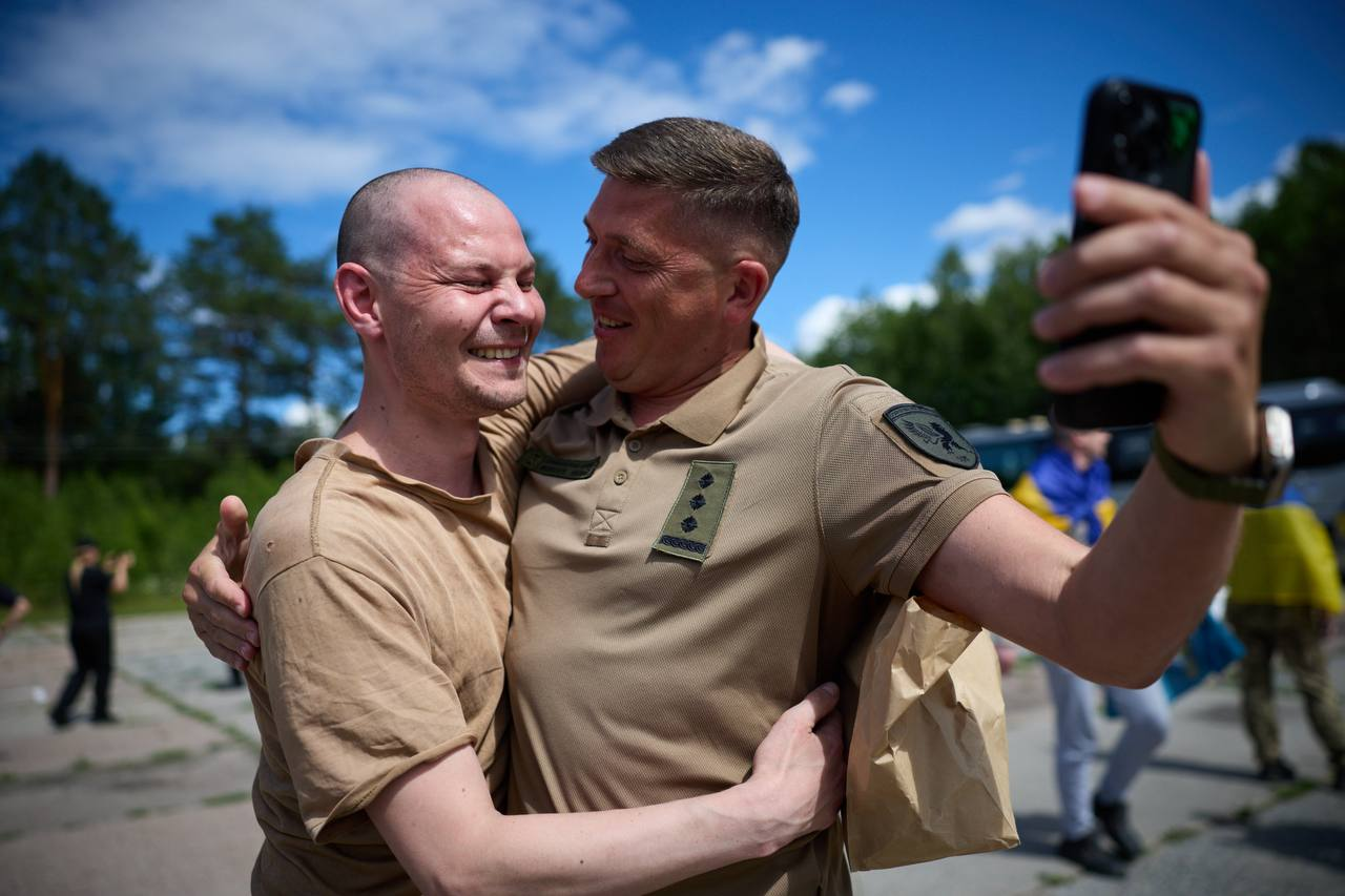 Двоє чоловіків радісно обіймаються просто неба, і один із них робить спільне селфі на смартфон. Чоловік праворуч одягнений у тактичну сорочку-поло пісочного кольору з погоном, а на розмитому задньому плані видно зелені дерева та блакитне небо з хмарами.