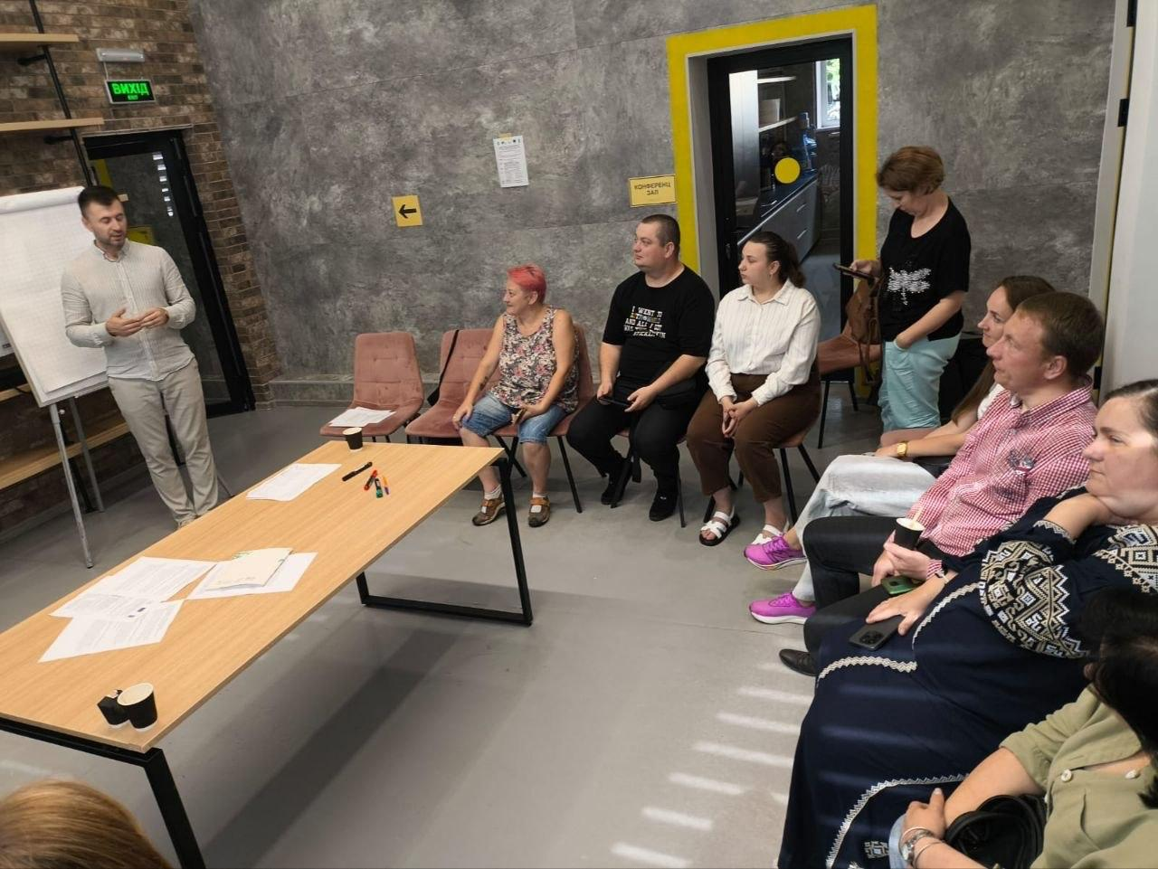 Фото з робочого воркшопу в конференц-залі. Ліворуч біля фліпчарта та столу з матеріалами стоїть ведучий, який спілкується з аудиторією. Справа на стільцях сидить група учасників, які слухають презентацію.