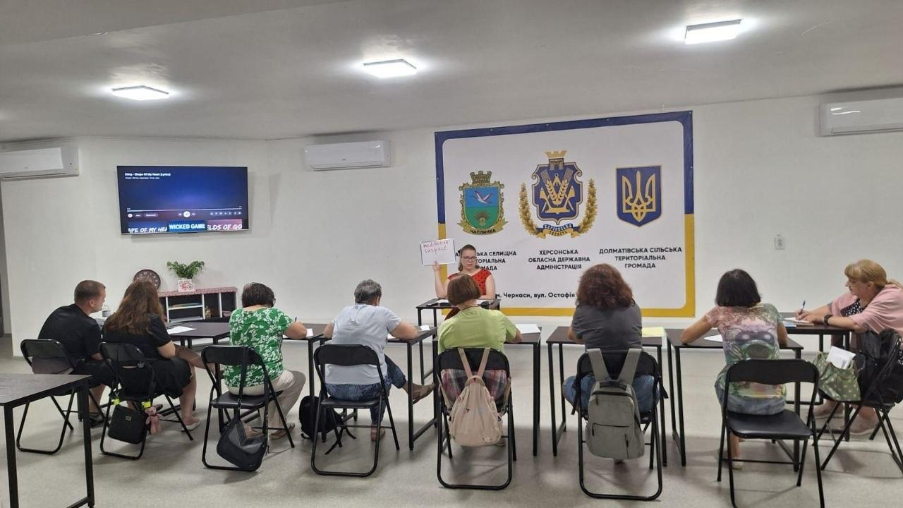 Фото з тренінгу в офіційному приміщенні з символікою Херсонської ОВА. Ведуча за столом демонструє плакат групі учасників, які сидять півколом і роблять записи.