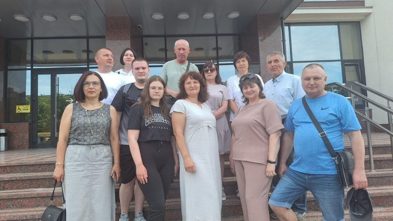 Групове фото учасників заходу на сходах біля входу до сучасної будівлі з великими вікнами та скляними дверима. Чоловіки та жінки різного віку стоять у кілька рядів і позирують для спільного знімка.