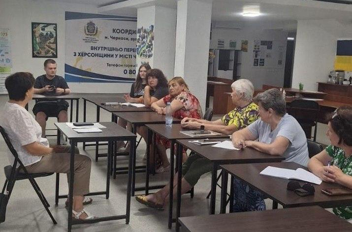 Фото консультації у Координаційному центрі для внутрішньо переміщених осіб з Херсонщини у місті Черкаси. Група людей сидить за столами, розставленими півколом, та спілкується з ведучою. На задньому плані розміщено інформаційний стенд із контактами гарячої лінії.