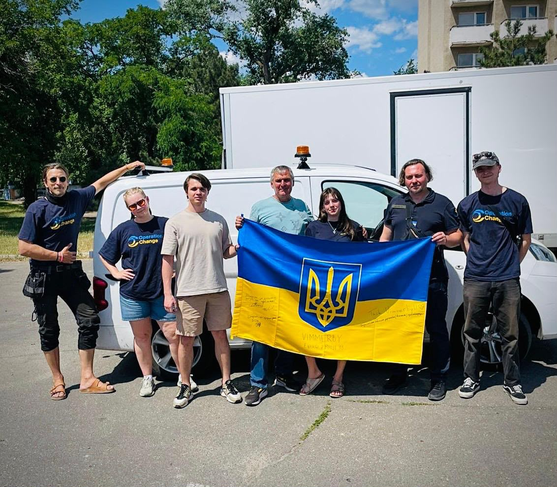 Групове фото волонтерів на вулиці біля білого вантажного автомобіля. Чоловіки та жінки тримають розгорнутий державний прапор України з гербом та підписами. Кілька учасників одягнені у брендовані футболки з логотипом організації.