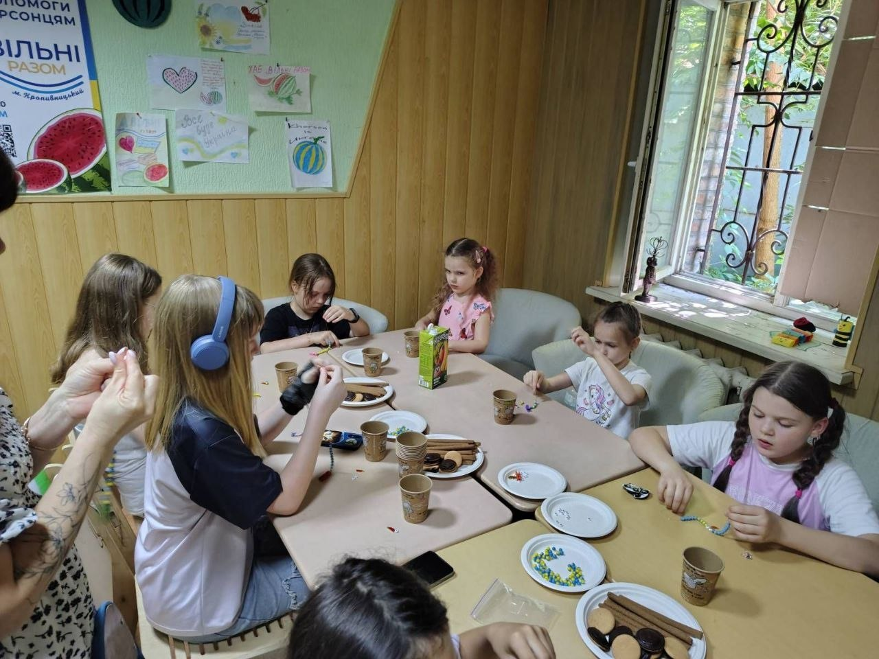 Фото з дитячого творчого майстер-класу в приміщенні хабу для переселенців «Вільні разом». Група дівчаток сидить навколо столів і займається рукоділлям із бісером та нитками. На задньому плані дерев'яна стіна прикрашена дитячими малюнками з кавунами та патріотичними написами.
