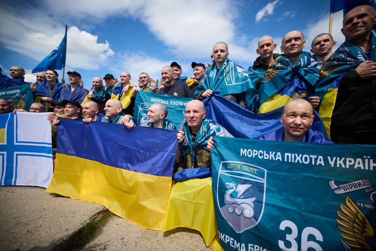 Групове фото звільнених з полону українських військовослужбовців просто неба. Чоловіки позирують для знімка, тримаючи розгорнуті державні прапори України та сині прапори Морської піхоти України, зокрема 36-ї окремої бригади морської піхоти.
