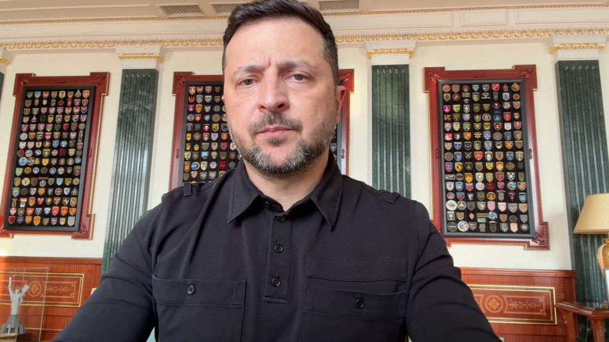 Чоловік у чорній сорочці з військовими кишенями дивиться в камеру на тлі офіційного кабінету. На стінах позаду нього висять стенди з багатьма різнокольоровими військовими шевронами.