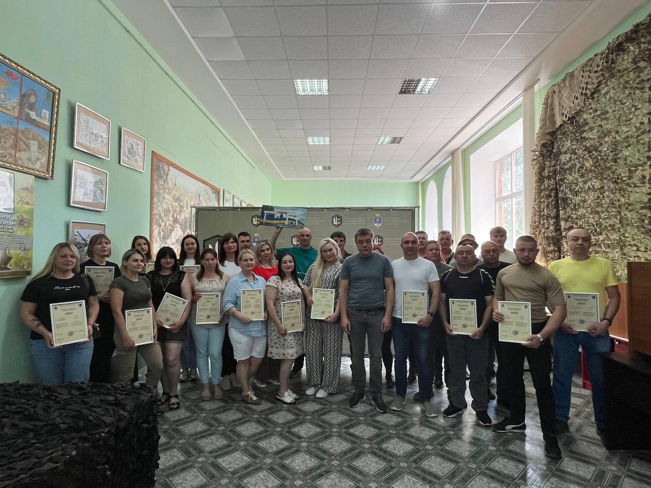 Групове фото людей у приміщенні з сертифікатами в руках, які стоять у два ряди на тлі стіни з картинами та вікнами з маскувальною сіткою.