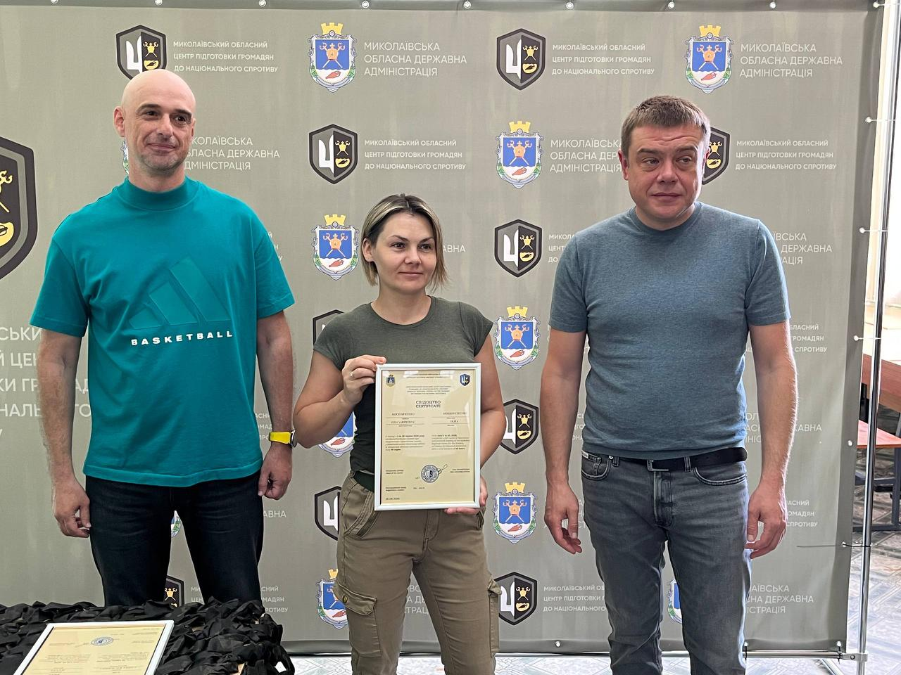 На знімку зафіксовано офіційне вручення сертифікатів випускникам навчального курсу. У центрі стоїть жінка у футболці кольору хакі з патріотичним принтом та тактичних штанах карго, яка тримає документ. Ліворуч від неї — чоловік у бірюзовій футболці «BASKETBALL» та чорних джинсах, а праворуч — чоловік у сірій футболці-поло та синіх джинсах. Захід проходить на урочистому тлі офіційного банера з логотипами організаторів.