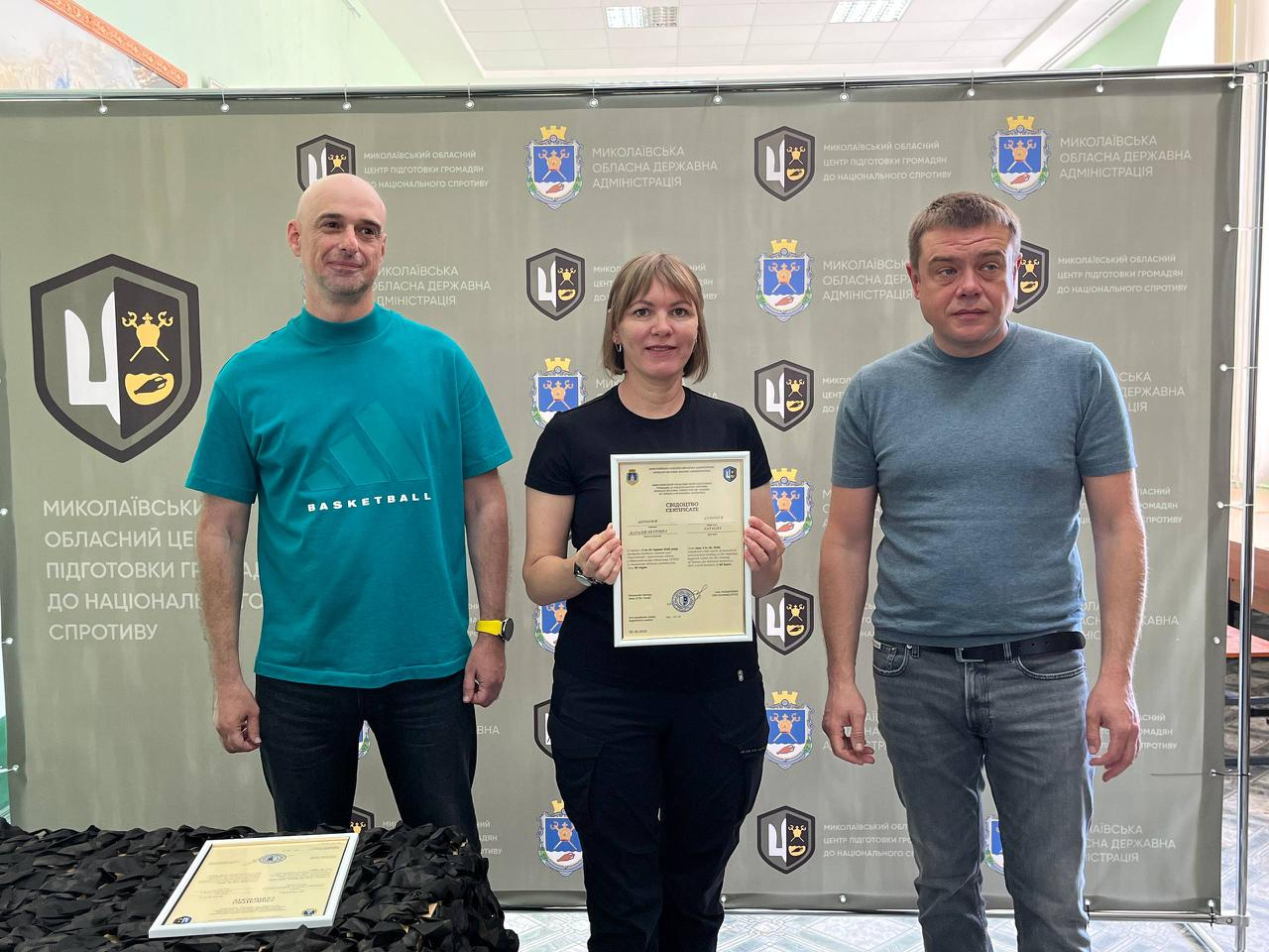 На знімку зафіксовано офіційне вручення сертифікатів випускникам навчального курсу. У центрі стоїть жінка у чорній футболці та темних штанах, яка тримає документ. Ліворуч від неї — чоловік у бірюзовій футболці «BASKETBALL» та чорних джинсах, а праворуч — чоловік у сірій футболці та синіх джинсах із ременем. Захід проходить на урочистому тлі офіційного банера з логотипами організаторів.