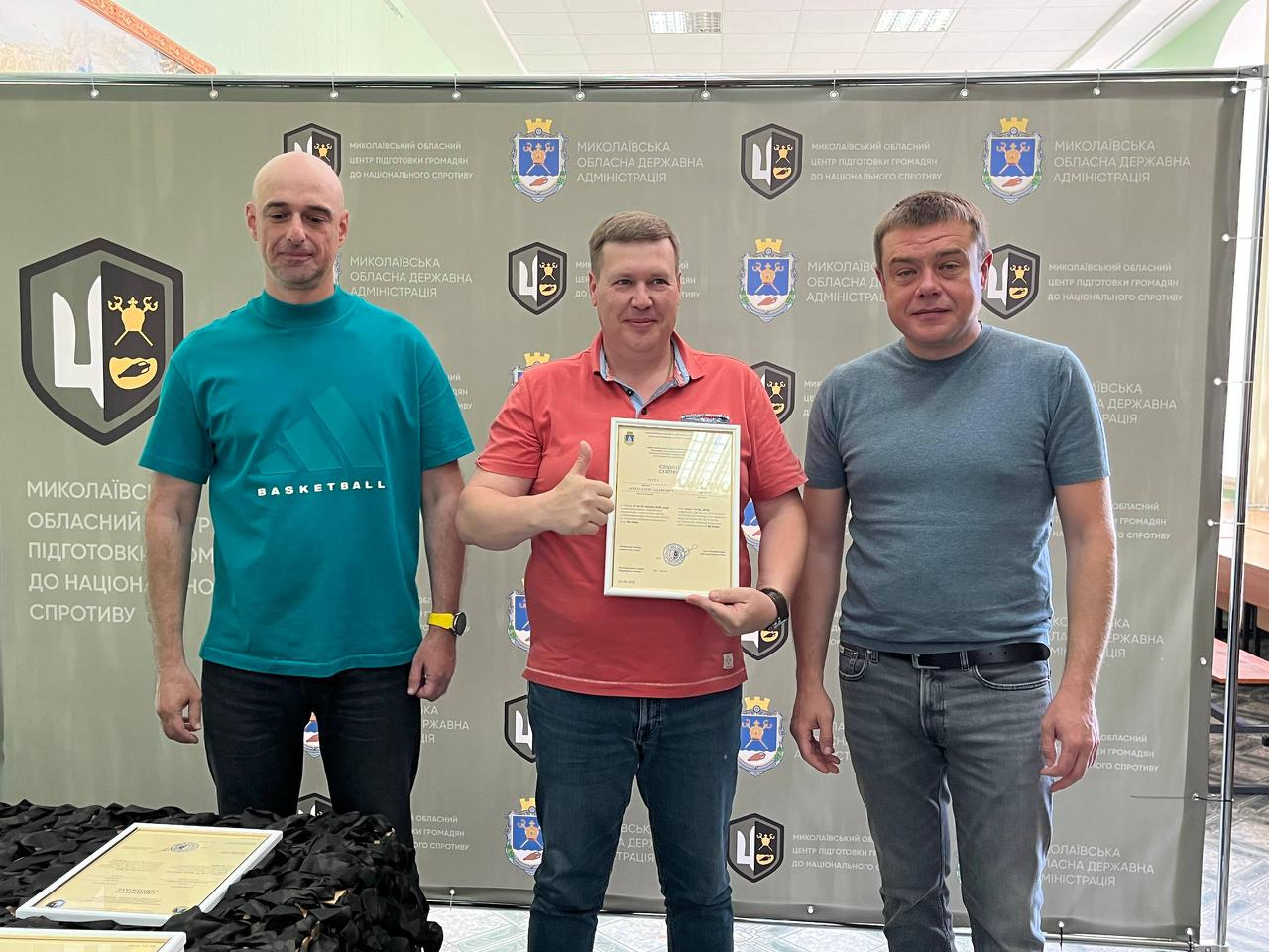 На знімку зафіксовано офіційне вручення сертифікатів випускникам навчального курсу. У центрі стоїть чоловік у червоній футболці-поло та синіх джинсах, який тримає документ і показує жест «великий палець угору». Ліворуч від нього — чоловік у бірюзовій футболці «BASKETBALL» та чорних джинсах, а праворуч — чоловік у сірій футболці та синіх джинсах із ременем. Захід проходить на урочистому тлі офіційного банера з логотипами організаторів.