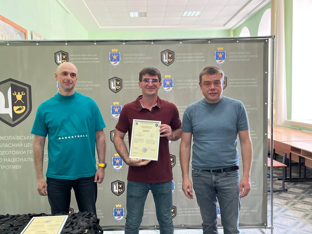 На знімку зафіксовано офіційне вручення сертифікатів випускникам навчального курсу. У центрі стоїть чоловік в окулярах, бордовій футболці-поло та синіх джинсах, який тримає документ. Ліворуч від нього — чоловік у бірюзовій футболці «BASKETBALL» та чорних джинсах, а праворуч — чоловік у сірій футболці та синіх джинсах із ременем. Захід проходить на урочистому тлі офіційного банера з логотипами організаторів.