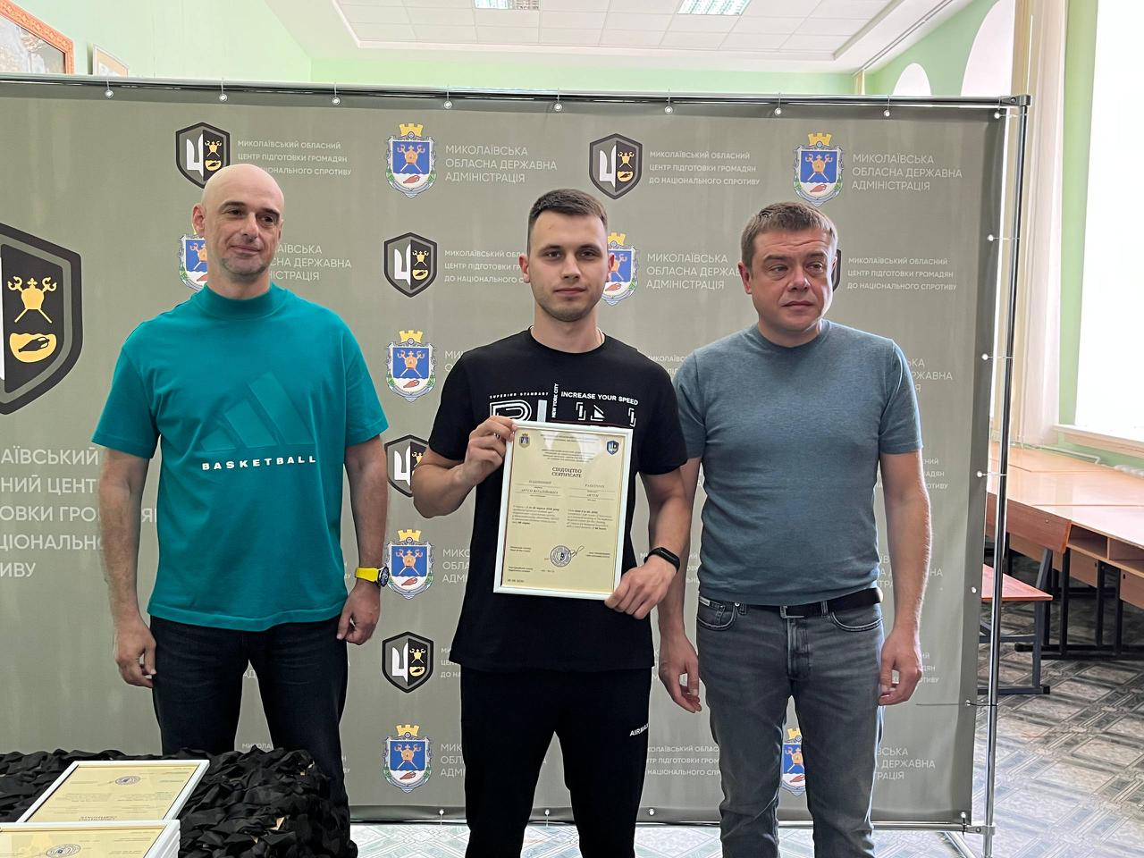 На знімку зафіксовано офіційне вручення сертифікатів випускникам навчального курсу. У центрі стоїть чоловік у чорній футболці з білим принтом та чорних спортивних штанах, який тримає документ. Ліворуч від нього — чоловік у бірюзовій футболці «BASKETBALL» та чорних джинсах, а праворуч — чоловік у сірій футболці та синіх джинсах із ременем. Захід проходить на урочистому тлі офіційного банера з логотипами організаторів.
