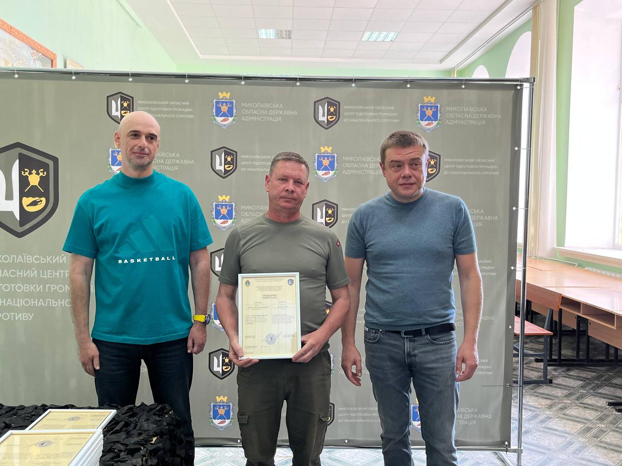 На знімку зафіксовано офіційне вручення сертифікатів випускникам навчального курсу. У центрі стоїть чоловік у футболці кольору хакі та темних штанах карго, який тримає документ. Ліворуч від нього — чоловік у бірюзовій футболці «BASKETBALL» та чорних джинсах, а праворуч — чоловік у сірій футболці та синіх джинсах із ременем. Захід проходить на урочистому тлі офіційного банера з логотипами організаторів.