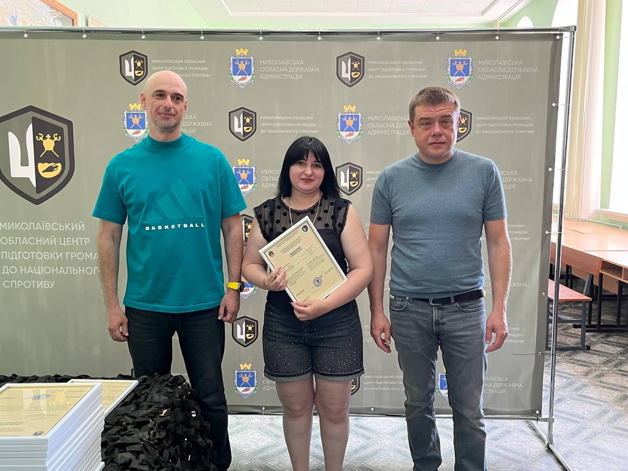 На знімку зафіксовано офіційне вручення сертифікатів випускникам навчального курсу. У центрі стоїть жінка у чорній блузці без рукавів з візерунком та темних шортах, яка тримає документ. Ліворуч від неї — чоловік у бірюзовій футболці «BASKETBALL» та чорних джинсах, а праворуч — чоловік у сірій футболці та синіх джинсах із ременем. Захід проходить на урочистому тлі офіційного банера з логотипами організаторів.