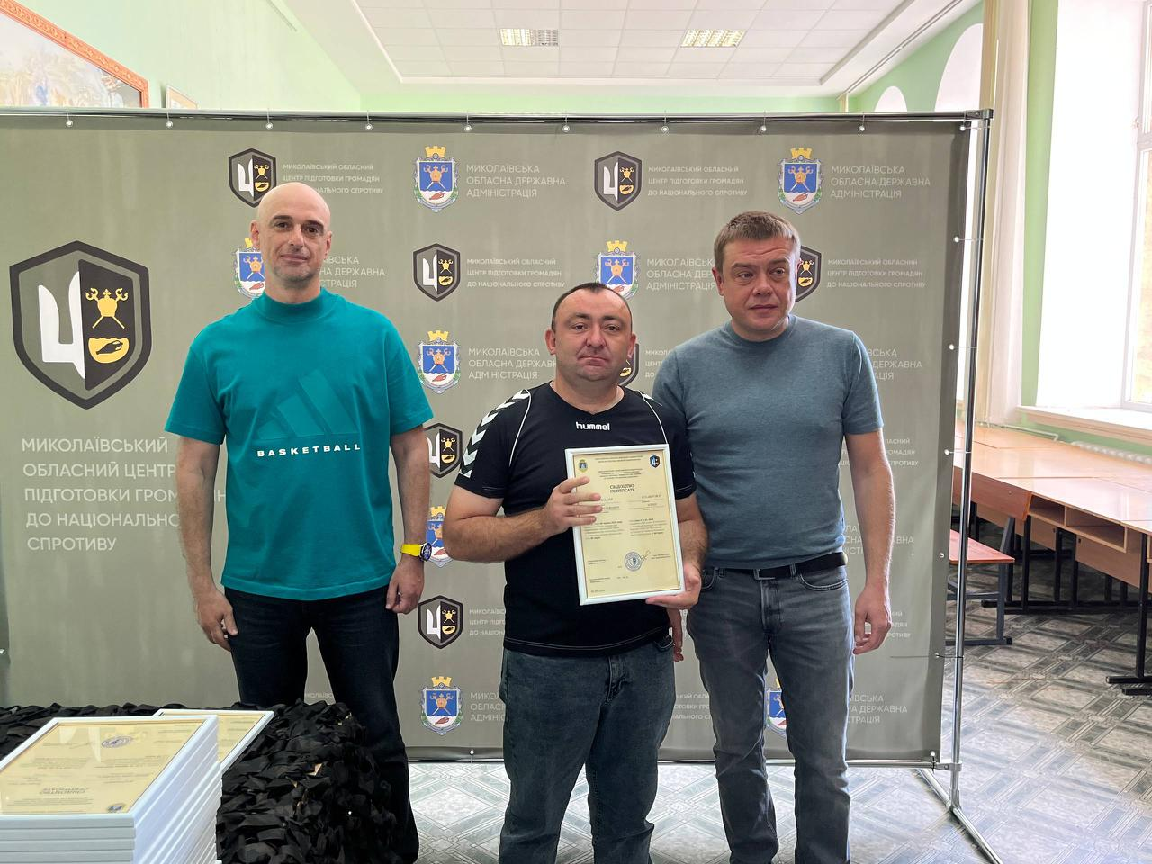 На знімку зафіксовано офіційне вручення сертифікатів випускникам навчального курсу. У центрі стоїть чоловік у чорній футболці «hummel» та темних джинсах, який тримає документ. Ліворуч від нього — чоловік у бірюзовій футболці «BASKETBALL» та чорних джинсах, а праворуч — чоловік у сірій футболці та синіх джинсах із ременем. Захід проходить на урочистому тлі офіційного банера з логотипами організаторів.