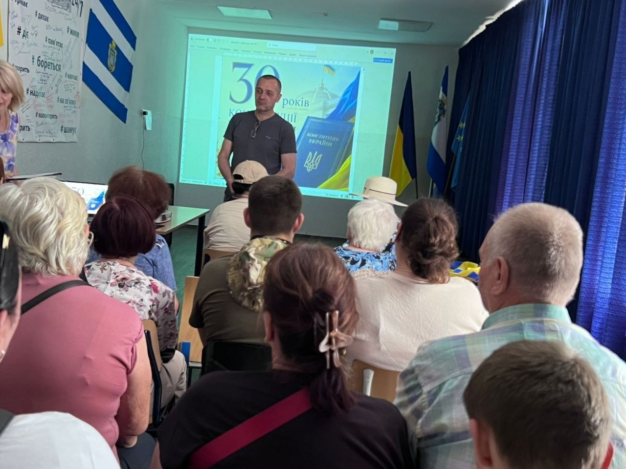 Чоловік виступає перед аудиторією у залі на тлі проєкційного екрана з презентацією до 30-річчя Конституції України та державних прапорів.