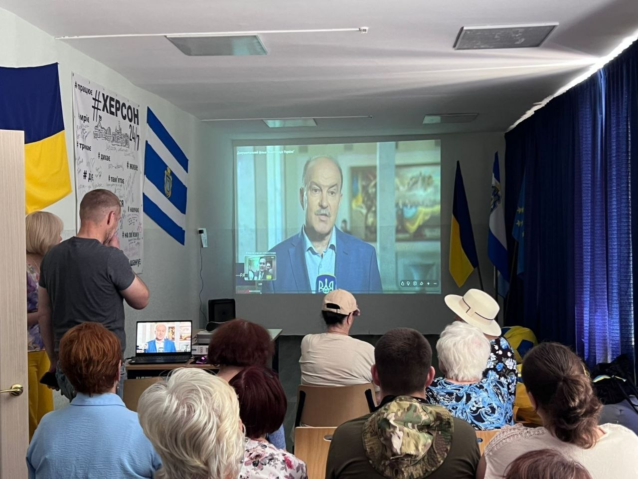 Аудиторія у залі слухає онлайн-виступ спікера, який транслюється на великому проєкційному екрані на тлі державних прапорів та патріотичного банера.