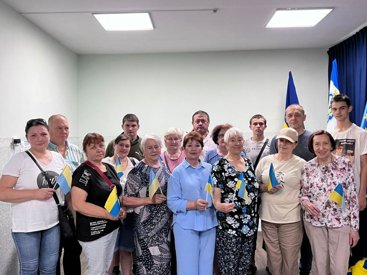 Групове фото людей різного віку у приміщенні, які стоять разом і тримають у руках маленькі прапорці України на тлі державних стягів.