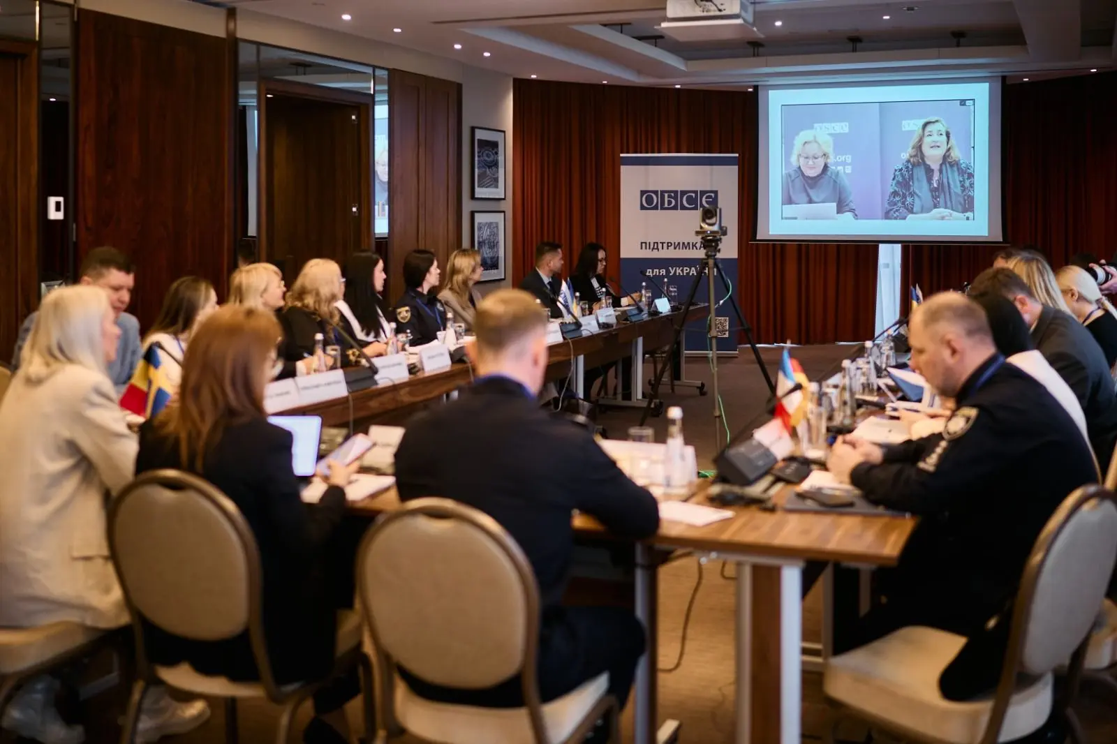 Учасники засідання сидять за великим Т-подібним столом у конференц-залі на тлі банера ОБСЄ та великого екрана з онлайн-трансляцією спікерів.