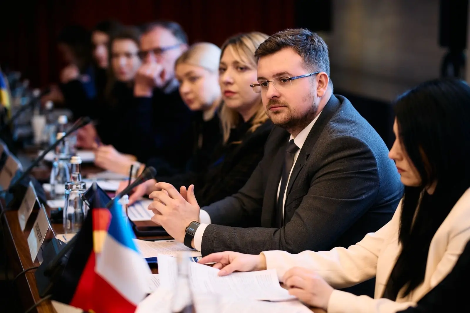 Учасники офіційного заходу сидять у ряд за столом засідань з документами та мікрофонами, уважно слухаючи спікера на тлі настільних наційональних прапорців.