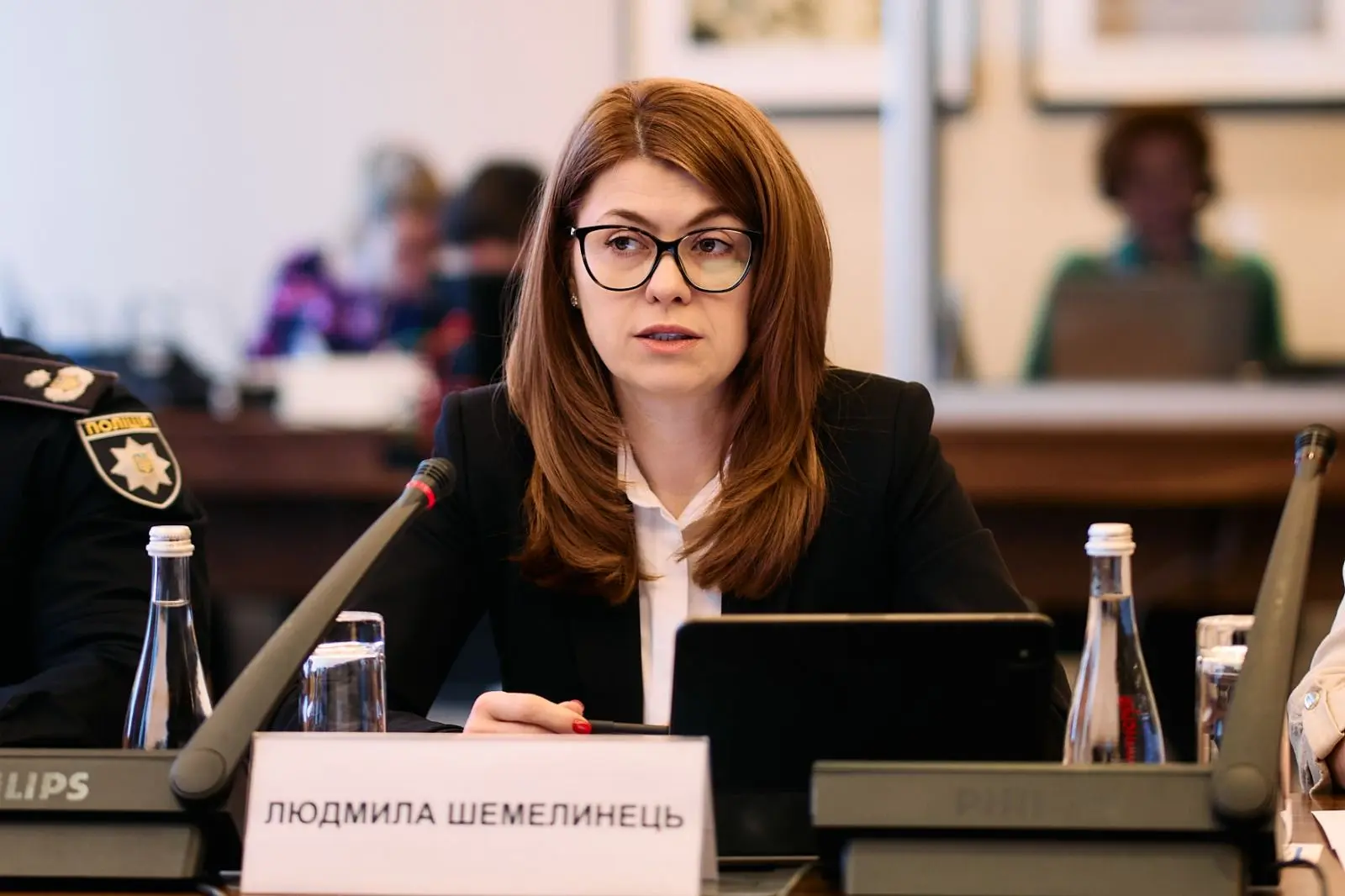 Жінка в окулярах та діловому костюмі сидить за столом засідань з мікрофоном, табличкою та планшетом на тлі інших учасників заходу, зокрема представника поліції.