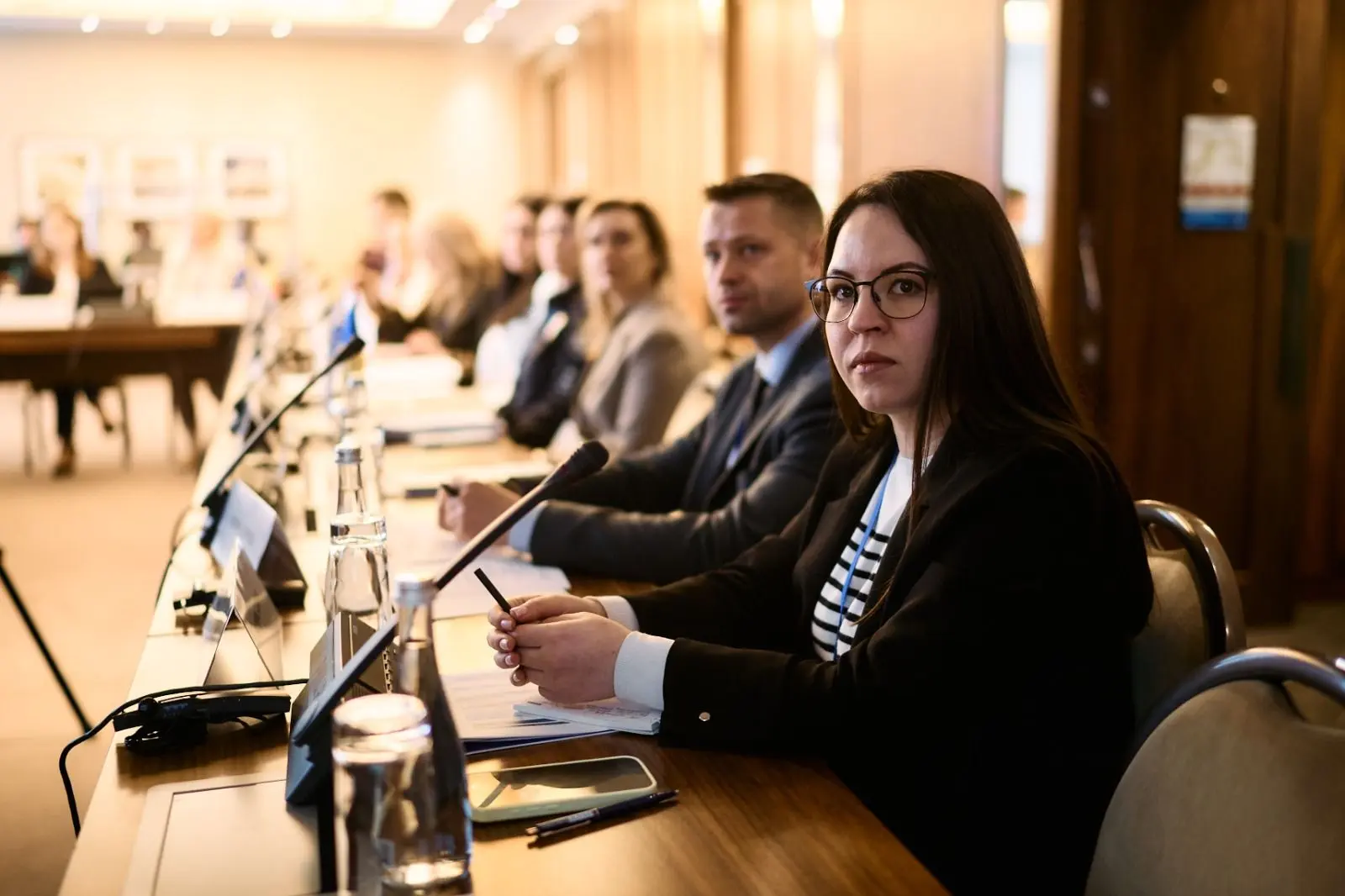 Жінка в окулярах та темному жакеті сидить на передньому плані за довгим столом засідань поруч з іншими учасниками офіційного заходу.