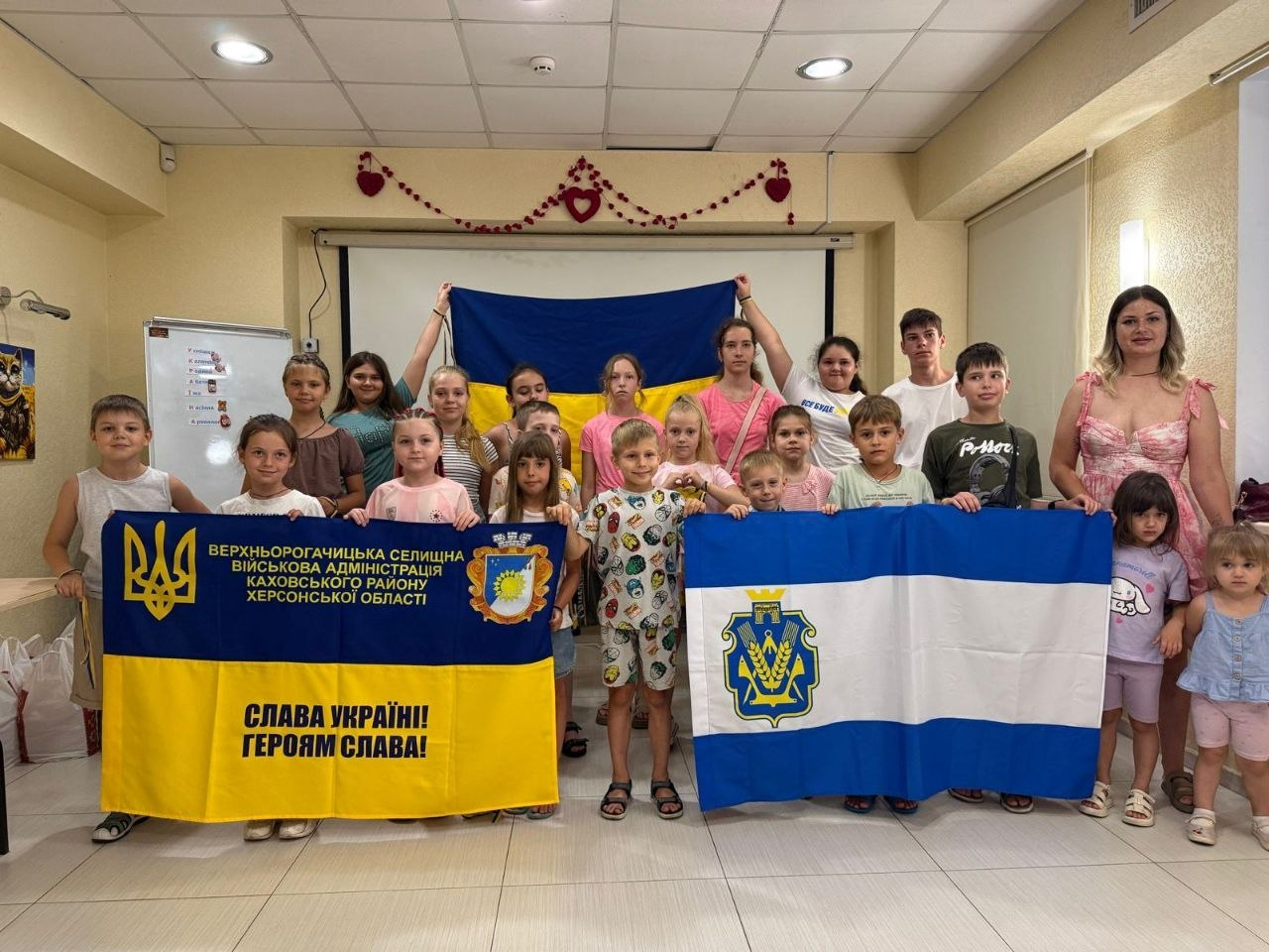 Група дітей та дорослих стоїть у приміщенні, тримаючи в руках державний прапор України, а також прапори Верхньорогачицької селищної військової адміністрації та Херсонської області.