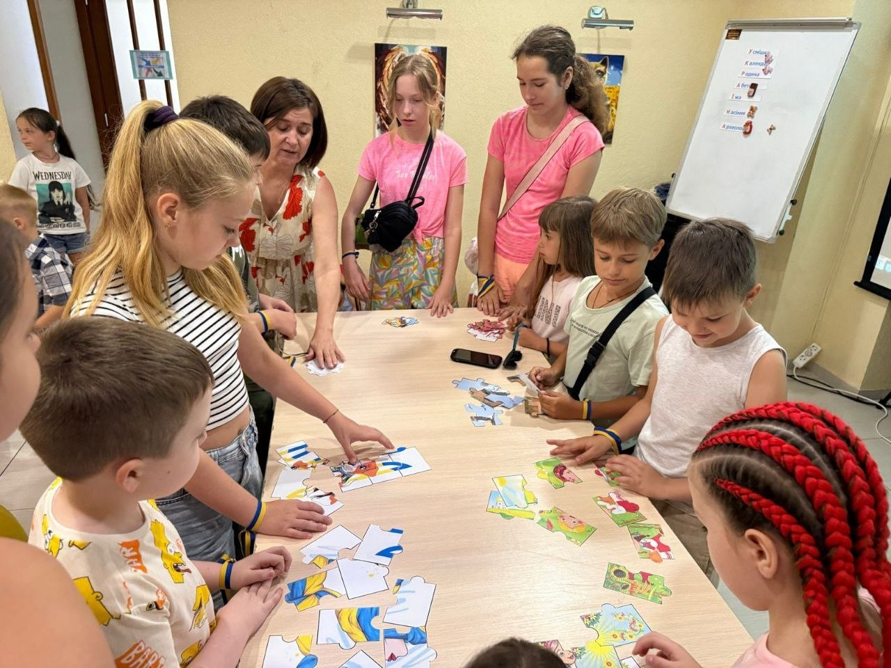 Діти різного віку разом із дорослою жінкою стоять навколо великого світлого столу в кімнаті та захоплено збирають різнокольорові пазли.