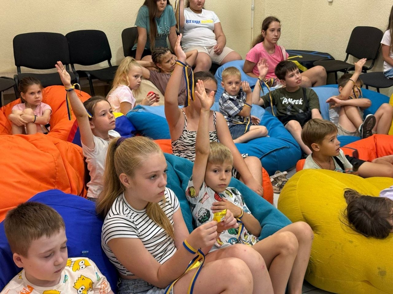 Діти та підлітки сидять у різнокольорових кріслах-мішках у приміщенні, уважно слухають і піднімають руки для відповіді, дехто має жовто-блакитні стрічки на зап'ястях.