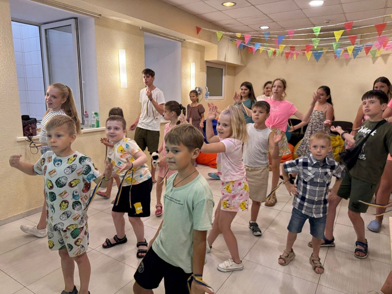 Діти та підлітки весело танцюють або повторюють рухи під час рухливої гри чи фізкультхвилинки у приміщенні, прикрашеному кольоровими прапорцями.