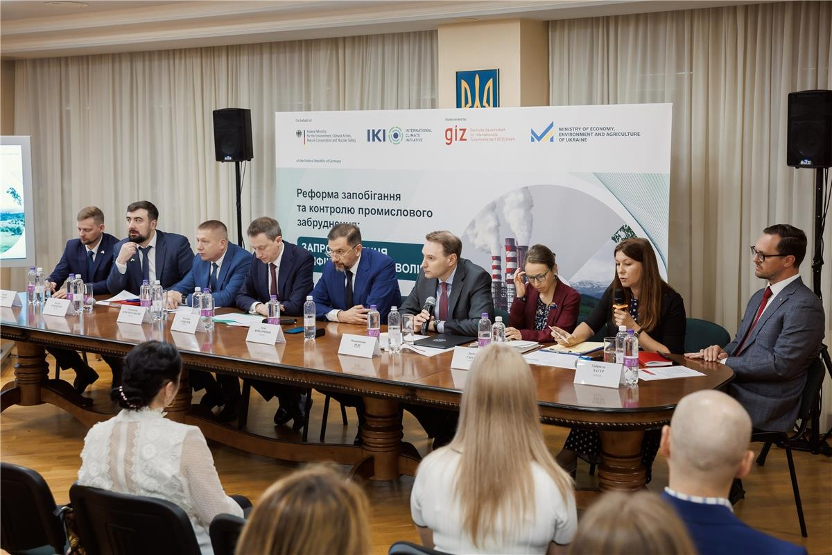 Учасники круглого столу сидять за великим дугоподібним дерев'яним столом у конференц-залі перед аудиторією на тлі офіційного банера, присвяченого реформі запобігання та контролю промислового забруднення, з логотипами партнерів, серед яких GIZ та Міністерство економіки.