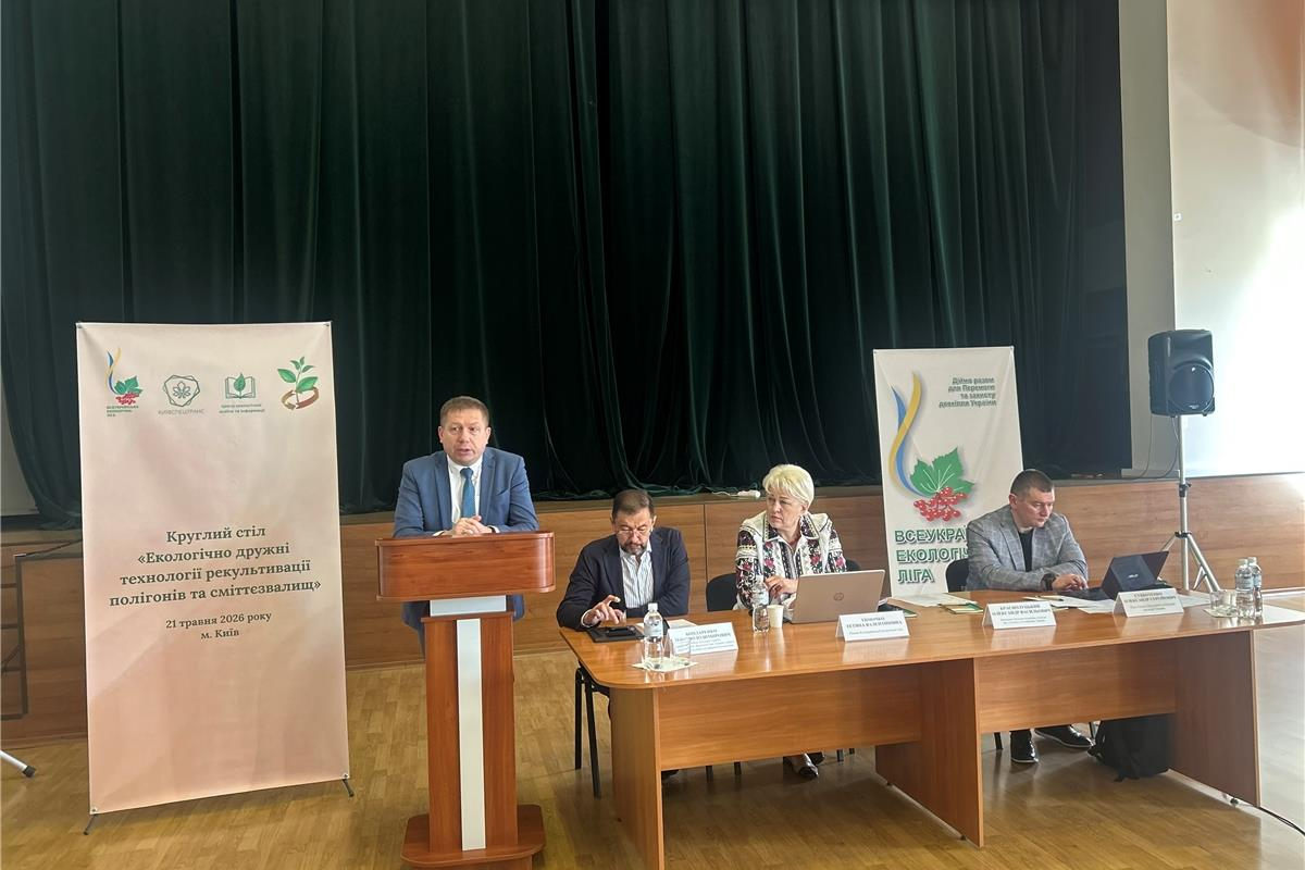 Спікер виступає за трибуною на офіційному заході, поруч із ним за дерев'яним столом сидять троє учасників, а на тлі темної завіси встановлено банери Всеукраїнської екологічної ліги та круглого столу про технології рекультивації полігонів.