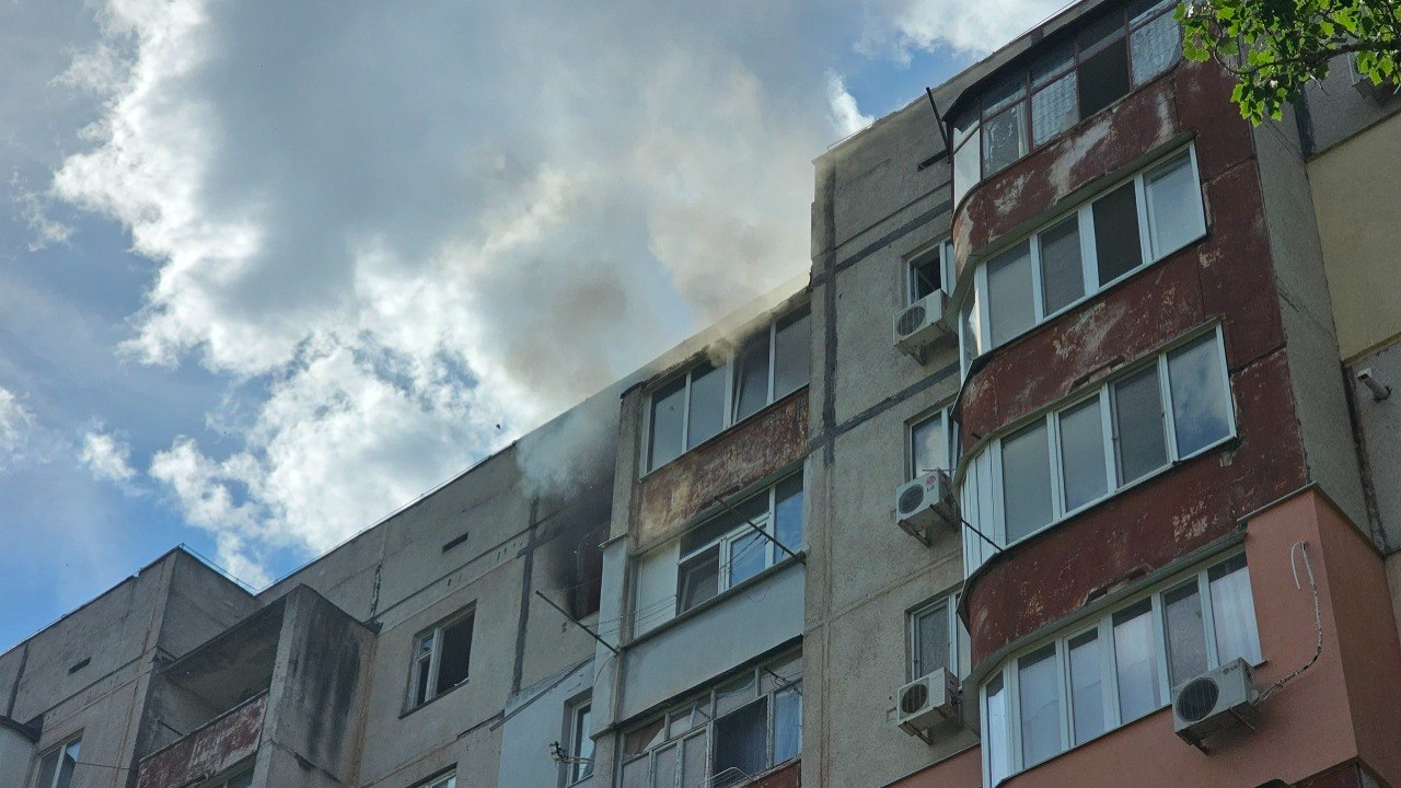 Багатоповерховий житловий будинок, з вікна одного з верхніх поверхів якого йде густий темний дим на тлі хмарного неба.