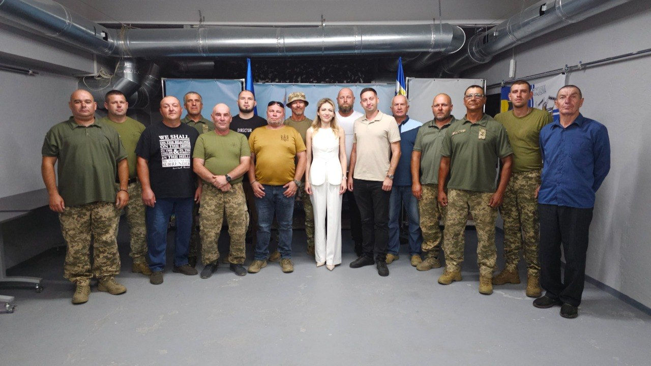 Групове фото людей у закритому приміщенні. У центрі стоїть жінка в білому костюмі, оточена групою з понад десяти чоловіків. Більшість чоловіків одягнені у військову форму або футболки захисного кольору. На задньому плані помітні державні прапори України.