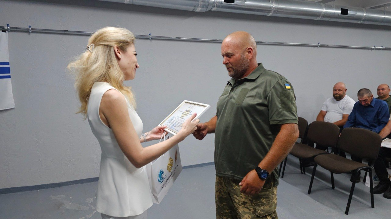 Жінка у білому костюмі вручає грамоту в рамці чоловікові міцної статури зі смуглим обличчям, який одягнений у тактичну сорочку кольору олива з нашивкою прапора України та камуфляжні штани. На задньому плані праворуч сидять інші люди.