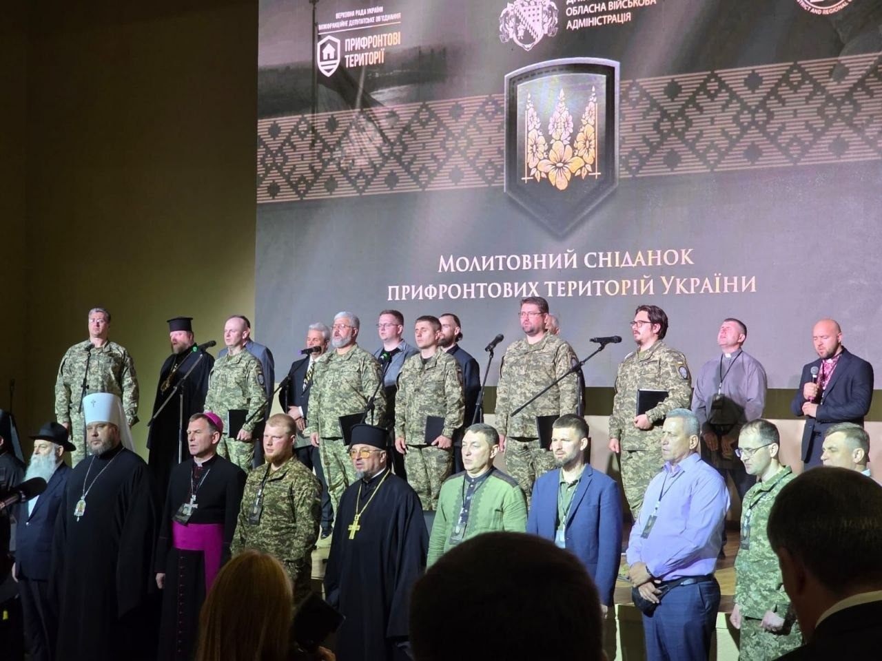 Група чоловіків у військовій формі, цивільному одязі та релігійному вбранні стоїть на сцені на тлі великого екрана з написом "Молитовний сніданок прифронтових територій України".