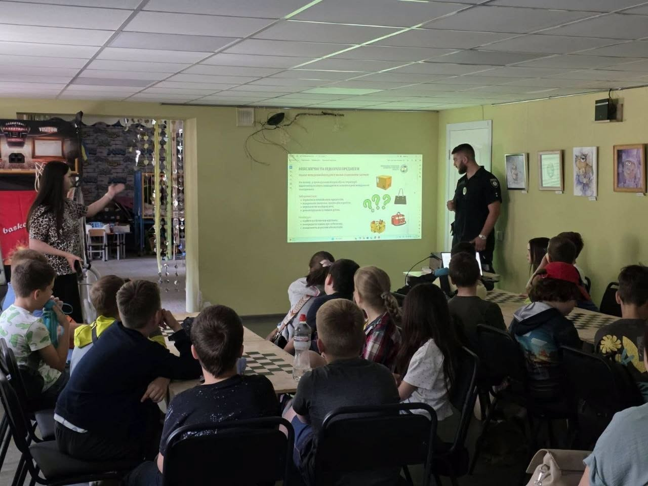 Група дітей сидить у класі та слухає лекцію, яку проводять двоє спікерів, зокрема чоловік у поліцейській формі, демонструючи презентацію на великому настінному екрані.