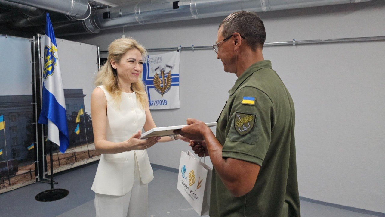 Жінка у білому костюмі вручає грамоту в рамці чоловікові в окулярах та військовій формі з нашивками на рукаві. Також вона тримає світлий паперовий пакет із написом «Kherson Region».