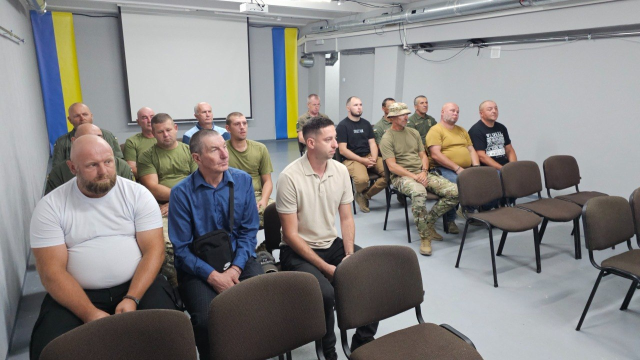 Група чоловіків сидить на стільцях у сірому залі, облаштованому під укриття, та слухає доповідача. Більшість присутніх одягнені у цивільний одяг або військову форму захисних кольорів. На задньому плані по боках висять синьо-жовті прапори.