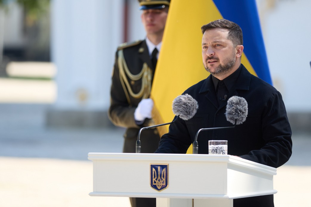 Чоловік у чорній сорочці та куртці виступає за білою трибуною з гербом України, на якій встановлено два мікрофони з вітрозахистом та склянку води. На задньому плані видно частину синьо-жовтого прапора та військовослужбовця почесної варти у парадній формі. Подія відбувається на вулиці за ясної сонячної погоди.