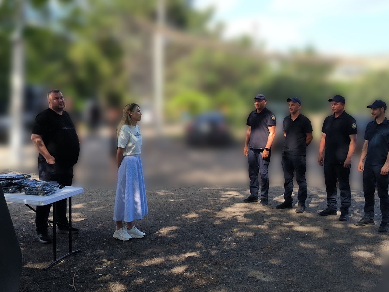 Жінка у білій футболці та довгій світло-блакитній спідниці виступає перед шеренгою з чотирьох чоловіків у темній форменому одязі та кепках. Ліворуч від неї стоїть чоловік у чорній футболці поруч із білим столом, на якому лежать речі в упаковках. Подія відбувається на вулиці, а задній план зображення сильно розмитий.